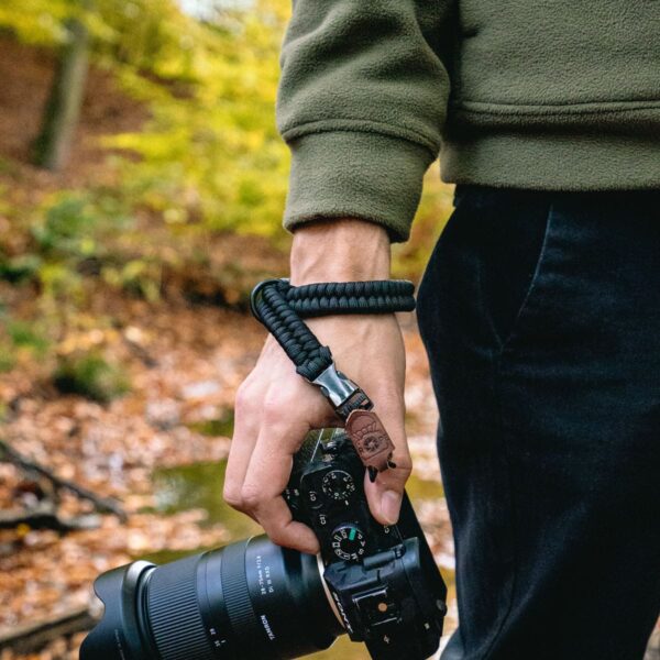 Das Bild zeigt eine Person im Wald, die ihre Kamera mit der C-Rope Handschlaufe The Claw in der Farbe Silent Black am Handgelenk gesichert hat.