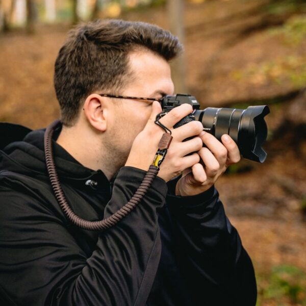 Hier zu sehen ist eine Person, die mit einer Kamera im Wald fotografiert und ihre Kamera mit dem Kamera Gurt The Traveler aus Paracord ind er Farbe Dark Coffee gesichert hat.