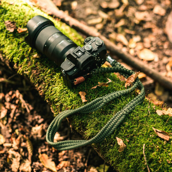 Abgebildet ist eine Kamera im Wald, an der der Kameragurt The Traveler aus Paracord in Military Olive befestigt ist.
