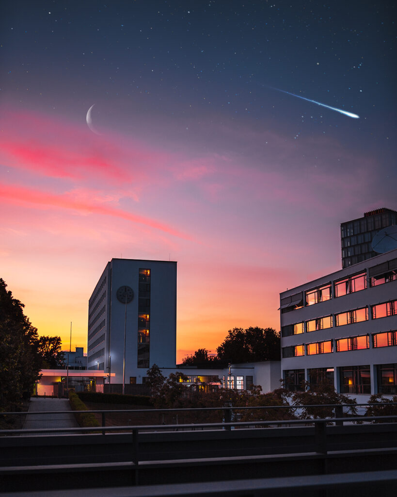 Das Bild zeigt Gebäude, bei einem verzaubernden Sonnenuntergang, mit Sternschnuppen und Mond am Himmel.