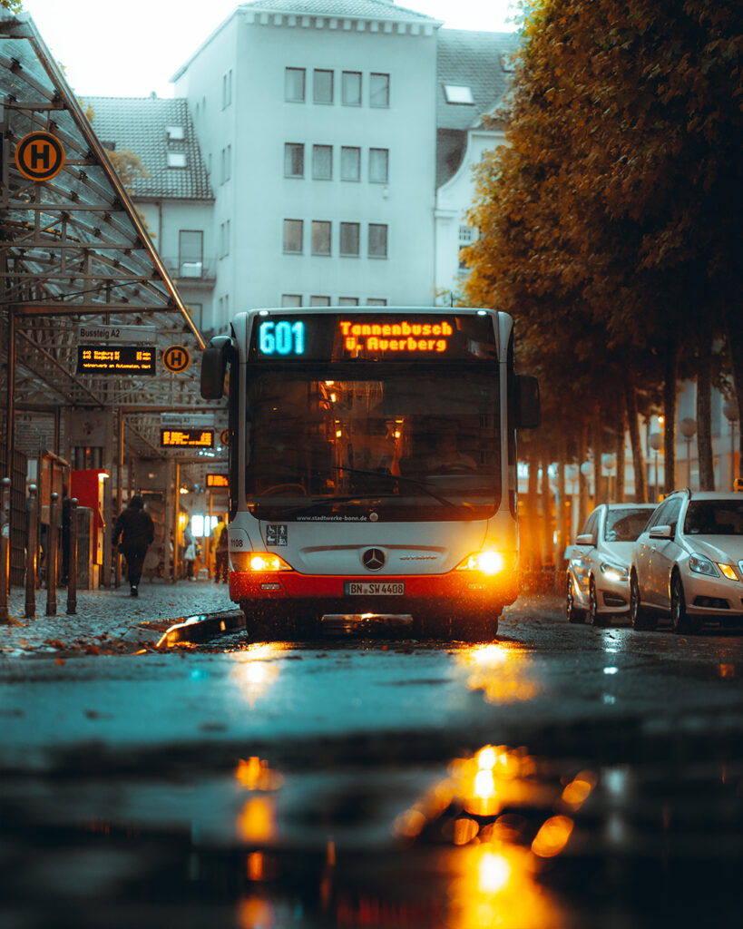 Das Bild zeigt einen Stadtbus, dessen Lichter sich in einer Regenpfütze spiegeln.