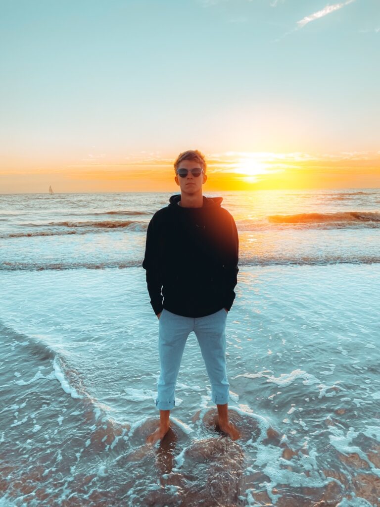 Auf dem Bild zu sehen ist Janik Over am Strand, wie er mit dem Rücken zum Sonnenuntergang, barfuß mit Sonnenbrille steht.