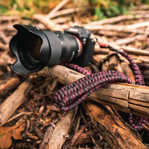 Abgebildet ist eine Kamera auf dem Waldboden, an der der C-Rope Kameragurt The Traveler in der Farbe Red Dots befestigt ist.