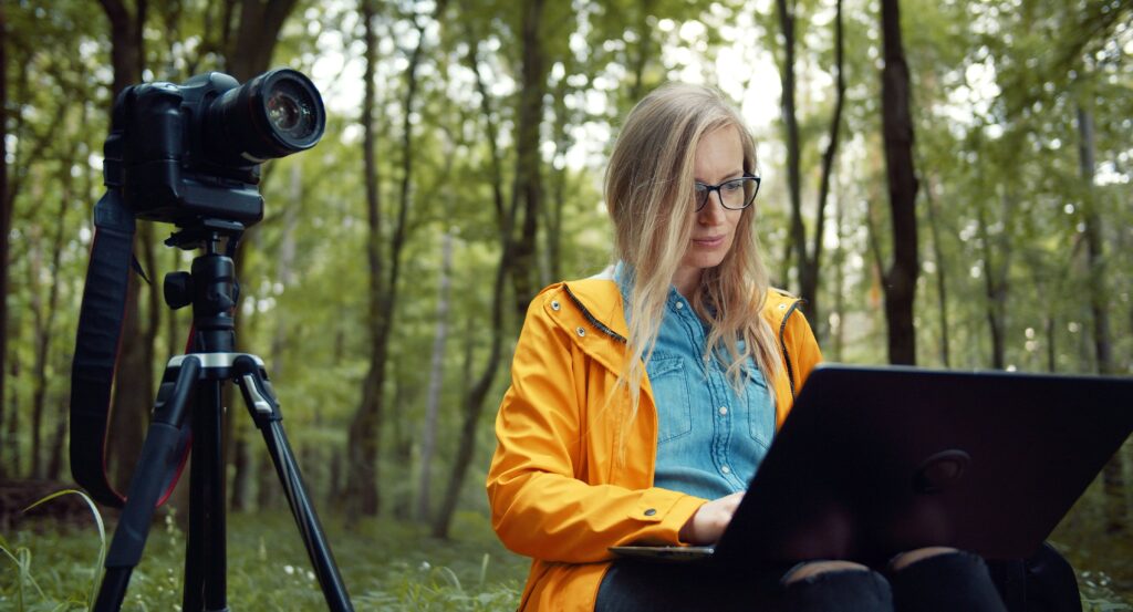Man sieht eine Frau im Wald, mit Kamera und Laptop.